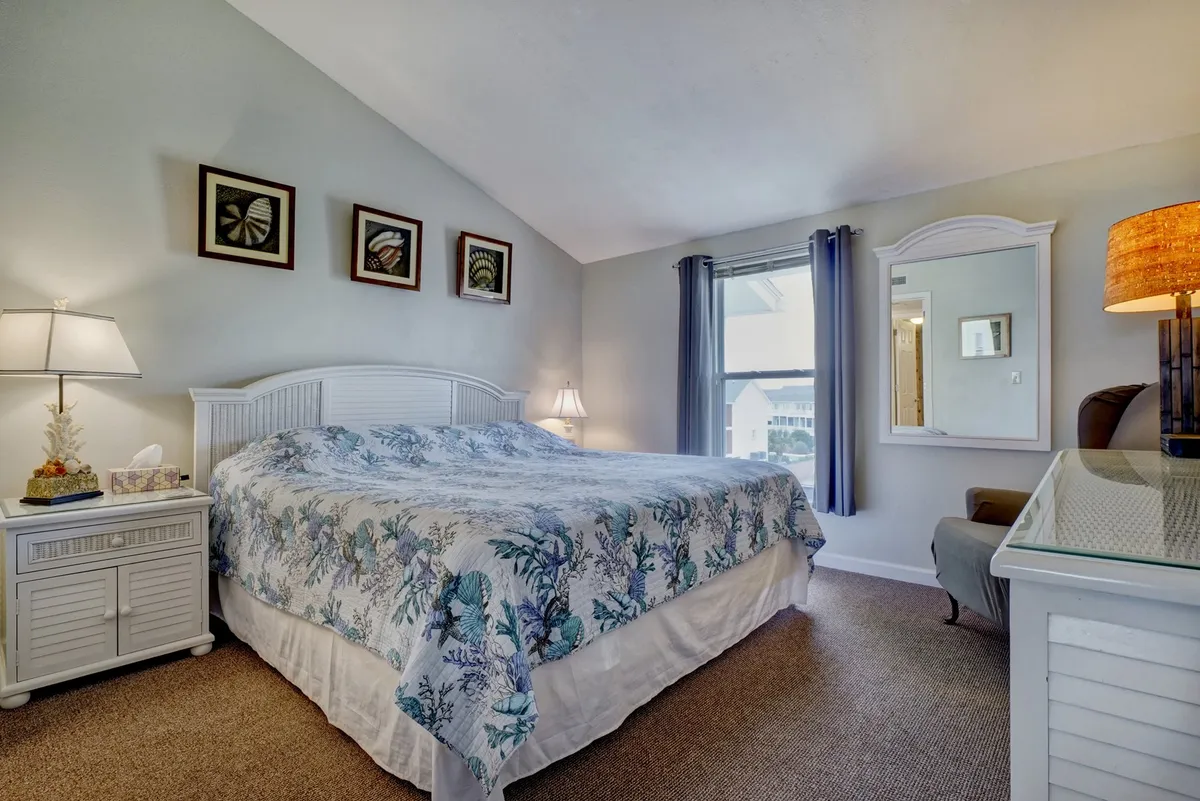 A cozy bedroom scene featuring a comfortable bed with soft bedding, decorative art on the walls, a bedside lamp, and a mirror. Soft natural light filters into the room, enhancing its restful and inviting atmosphere.