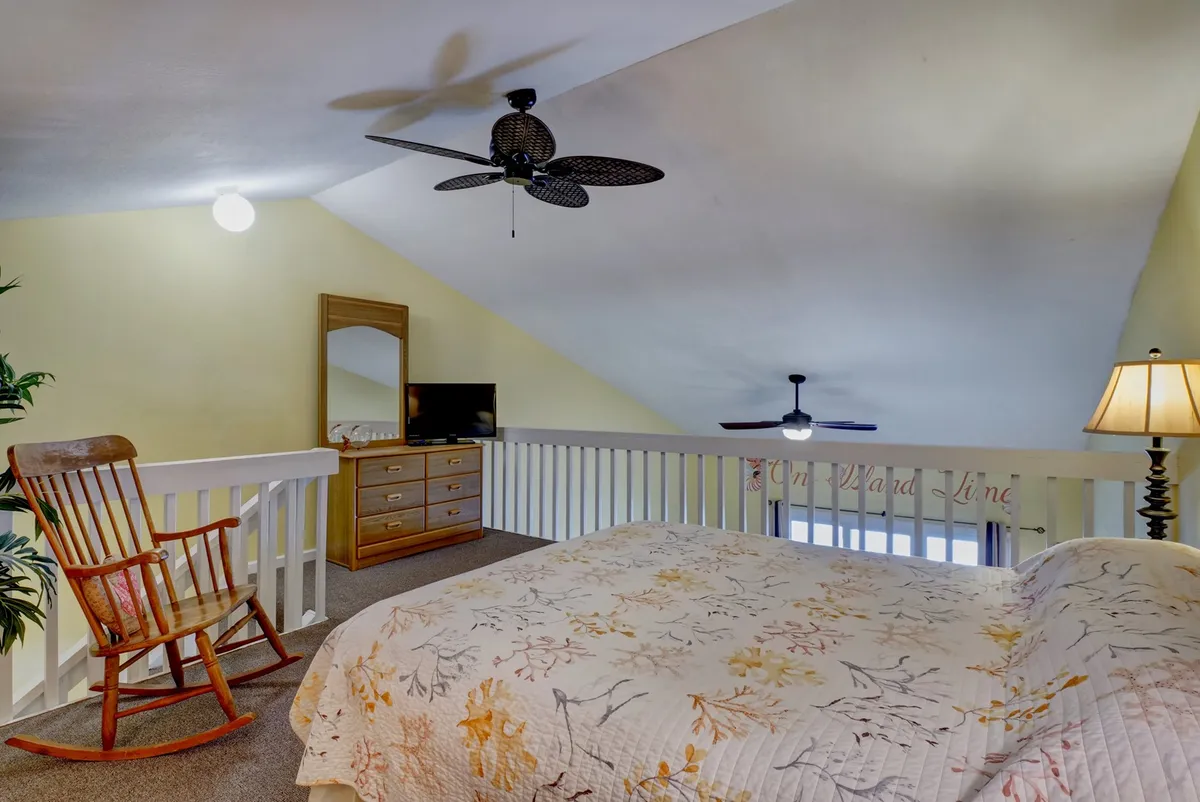 A loft area featuring a bed with a floral quilt, a classic rocking chair, wooden dresser, TV, and soft lighting. The elevated vantage point hints at views and a sense of privacy, offering a relaxed, inviting vibe.
