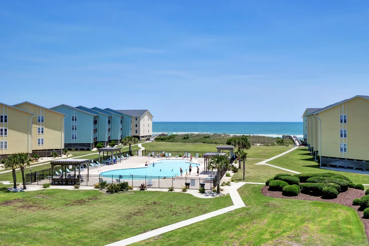 This vibrant exterior shot centers on the sparkling swimming pool surrounded by lush landscaped gardens and Surf Condos buildings. Several people are enjoying the pool and lounging nearby, conveying an inviting, lively atmosphere. The ocean is visible in the background, tying in the coastal vibe.