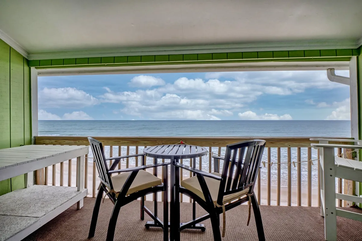 The image showcases a serene interior space with green accent walls, cozy outdoor seating on a private balcony, and a captivating, unobstructed ocean view. The setting hints at tranquil activities like enjoying coffee at sunrise, reading a book, sharing meals, or savoring sunsets together.