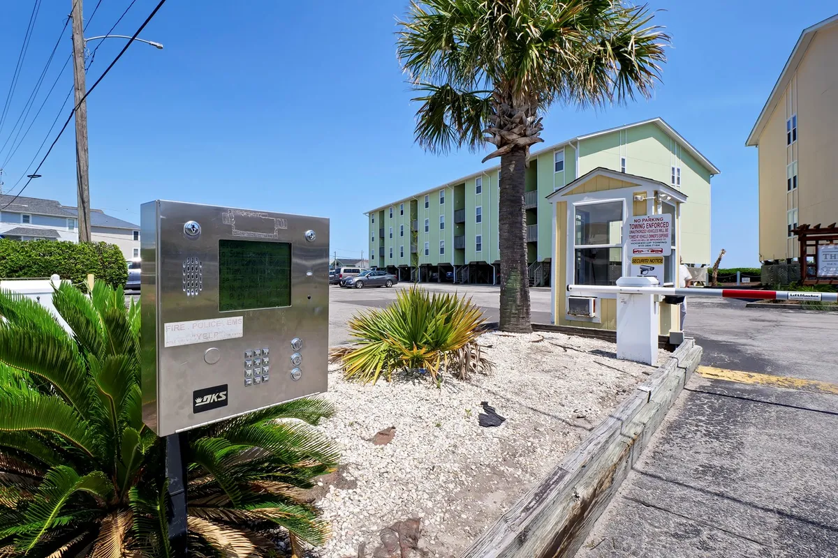 This exterior image features the entrance area of Surf Condos, with palm trees, green buildings, an intercom system, and a welcoming kiosk. The landscaping and relaxed mood set the scene for arrivals and departures, offering context for the guest experience upon arrival.