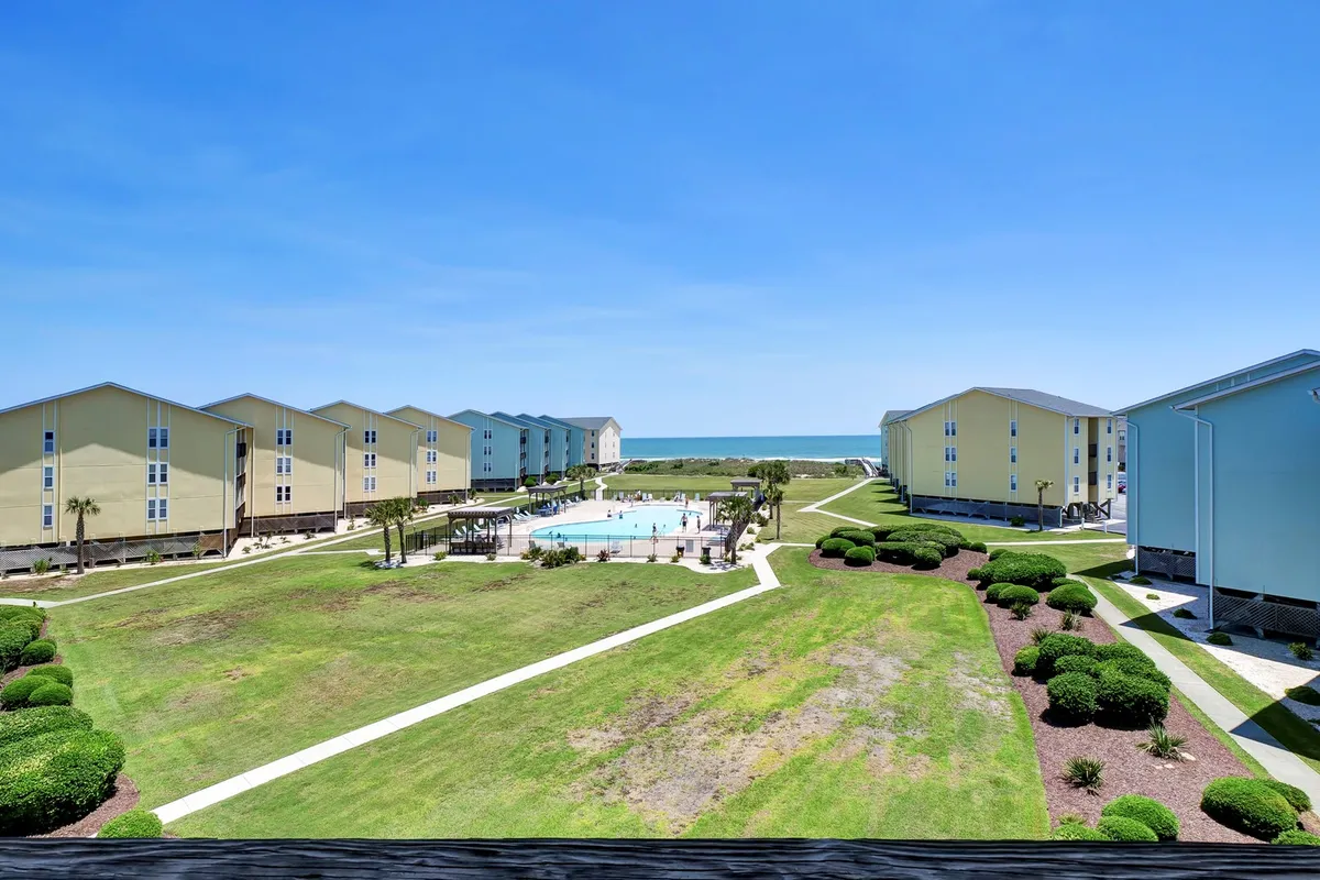 An inviting swimming pool with a green lawn and ocean views, surrounded by the complex's buildings. Sunbathers, swimmers, and families enjoying picnics are visible, all basking in a relaxed coastal atmosphere.