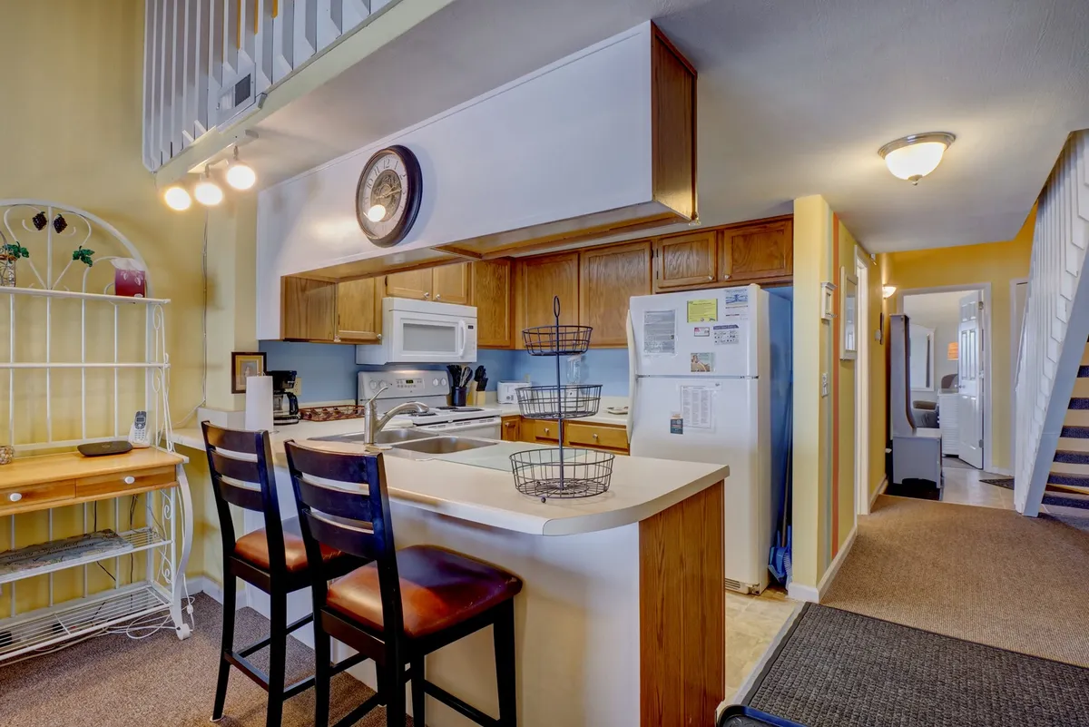 This image showcases a cozy, functional kitchen with an island, open layout, wooden cabinets, a wall clock, and welcoming bar stools. The scene hints at a space designed for both cooking and socializing, making it easy for multiple people to gather and prepare food together.
