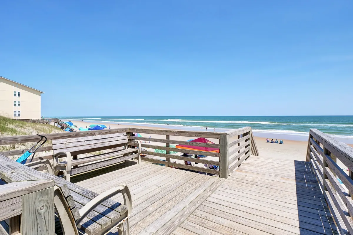 A broad view from the deck looking out over the sandy beach, with beach umbrellas, sunbathers, and the sparkling ocean stretching into the distance. The image captures the sense of place and easy access to the seaside.
