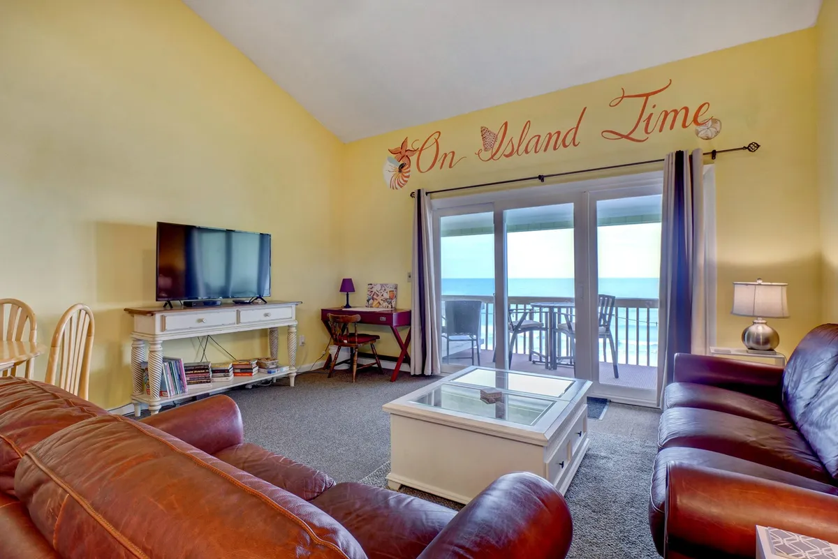 This image features a spacious living area centered around a plush sofa set, with decorative marine-inspired wall art. Large sliding glass doors lead outside to a deck, inviting in ample sunlight and offering an immediate visual connection to the outdoors.